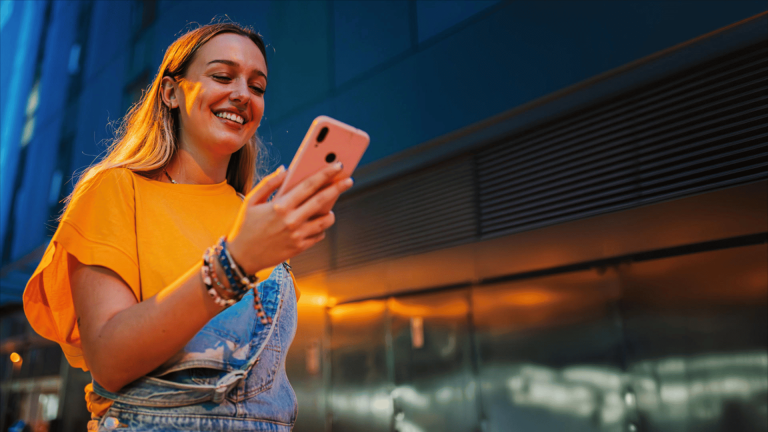 Mujer joven usando su celular con una sonrisa mientras camina por la ciudad al atardecer